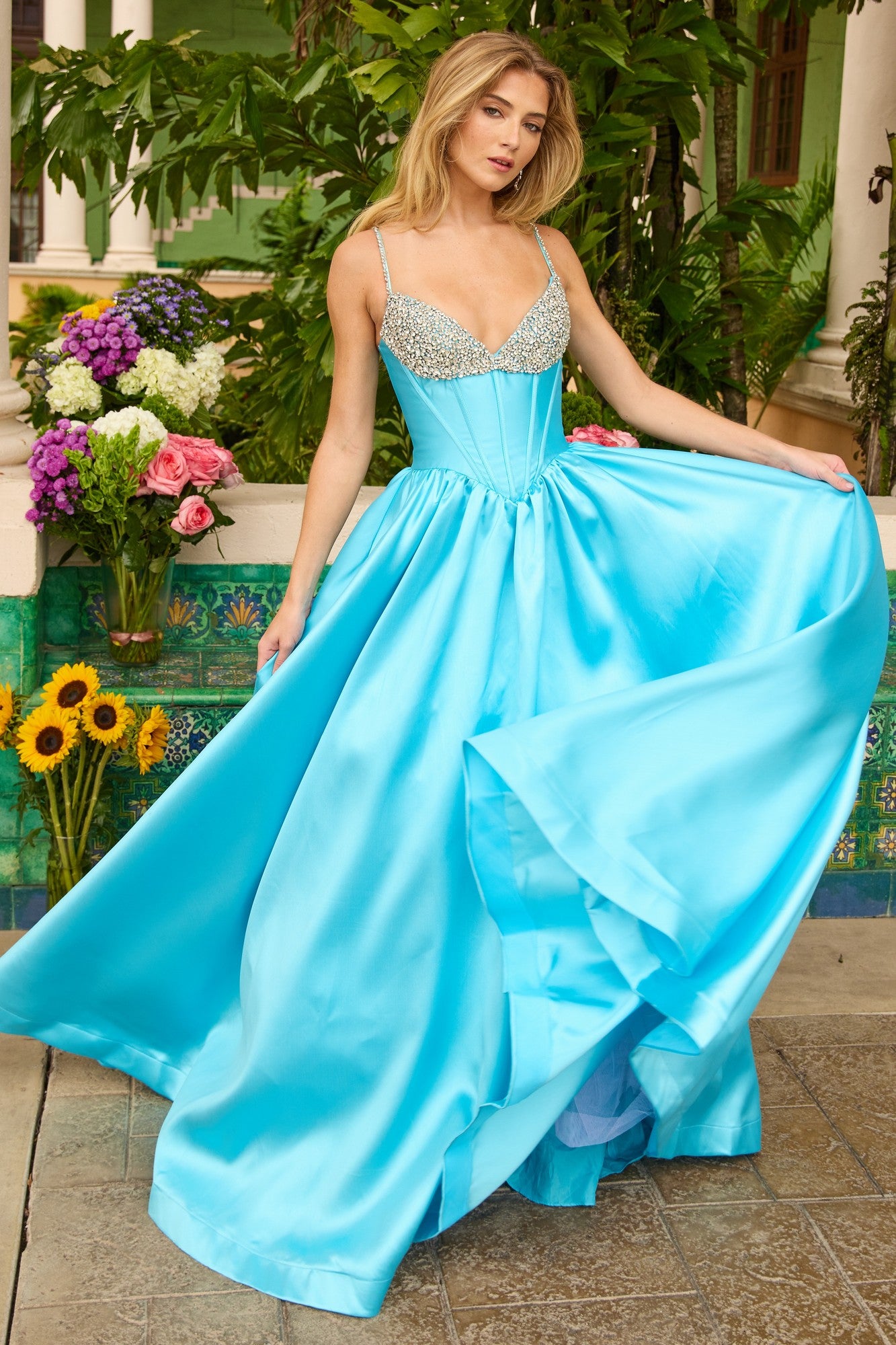 A woman poses outdoors near flowers and greenery wearing the Ava Presley 42311 long prom dress in turquoise, featuring a silver embellished corset bodice.